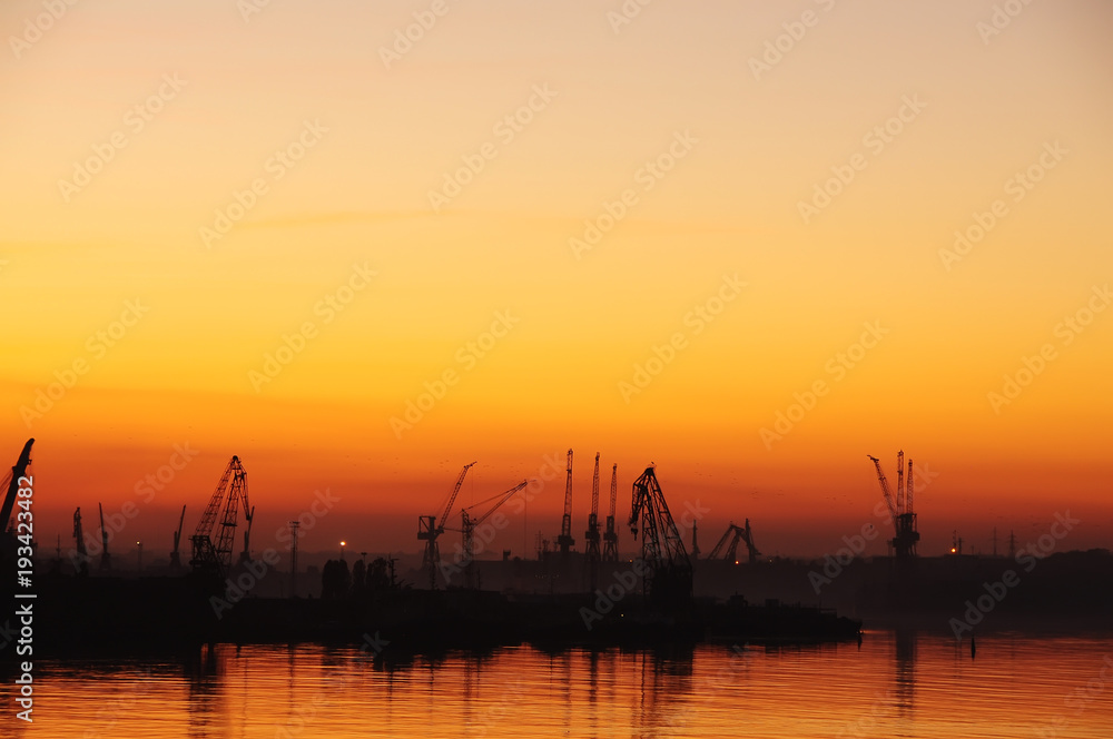 Silhouettes of cranes in the port and reflection in the water at sunset. Dark photo.
