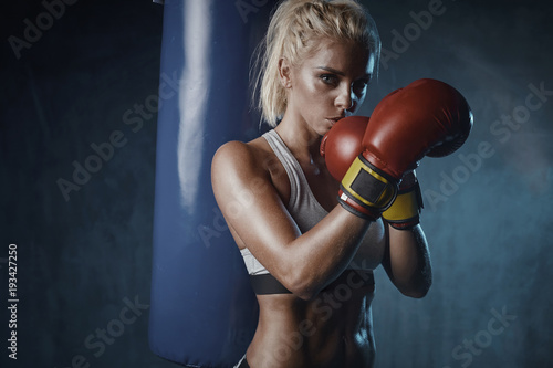 Portrait of sexy blonde boxer woman in red boxing gloves and grey sports bra  standing near blue punching bag with hands crossed near face