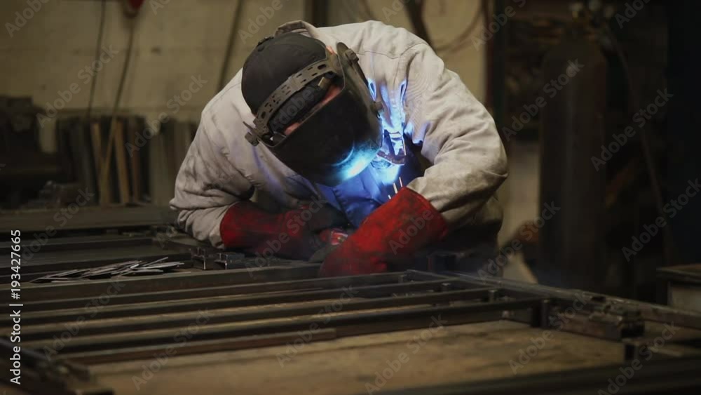 Man is wearing welding helmet and fireproof red gloves, conducting welding works. He is standing near workplace in a workshop Man is wearing welding helmet and fireproof red gloves, conducting welding works. He is standing near workplace in a workshop