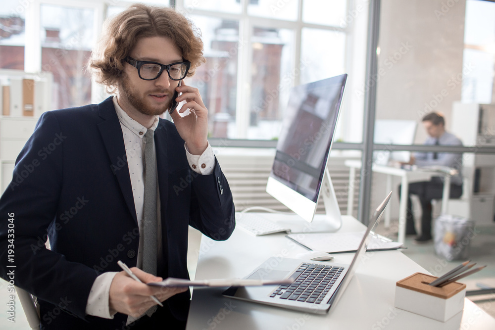 Busy office worker in formalwear talking by phone and reading papers by workplace Busy office worker in formalwear talking by phone and reading papers by workplace