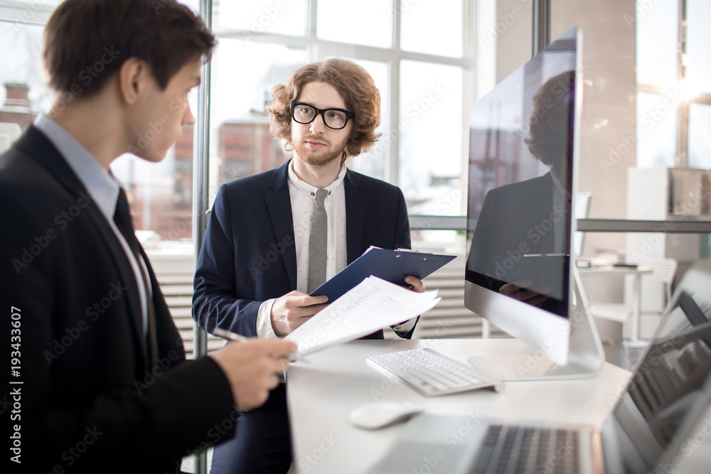 Two agents or bankers with documents sitting byb desk in office and discussing financial information Two agents or bankers with documents sitting byb desk in office and discussing financial information