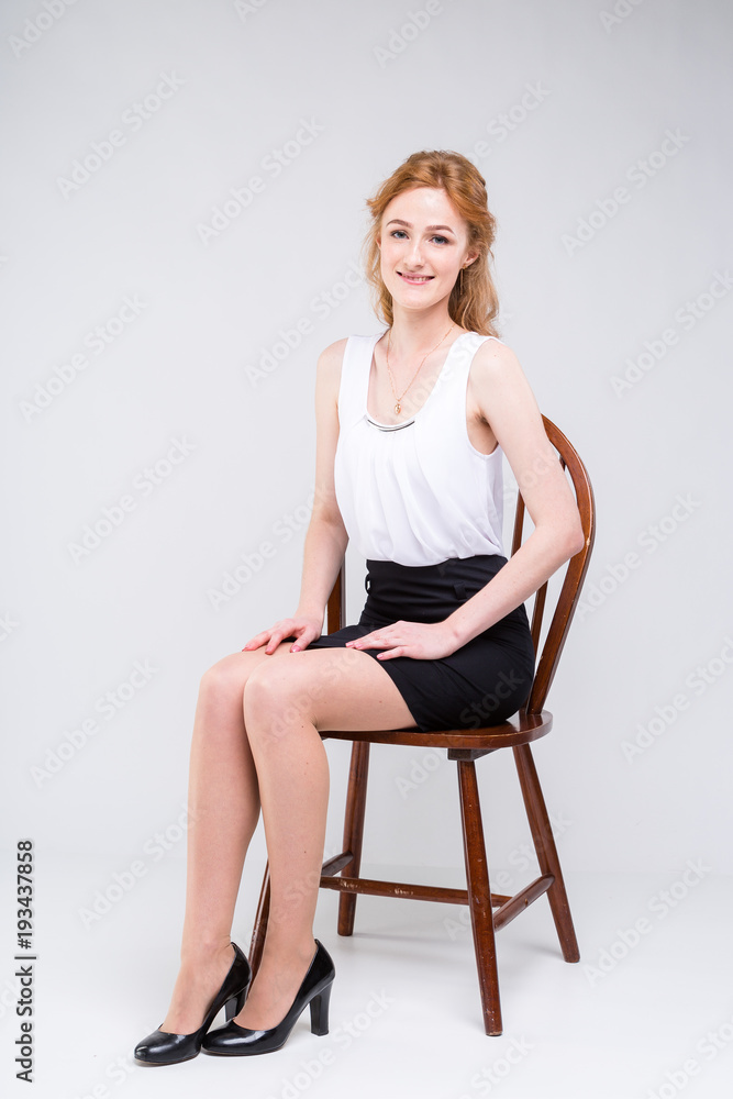 Portrait of beautiful business woman with long red, curly hair sitting on  wooden chair on white