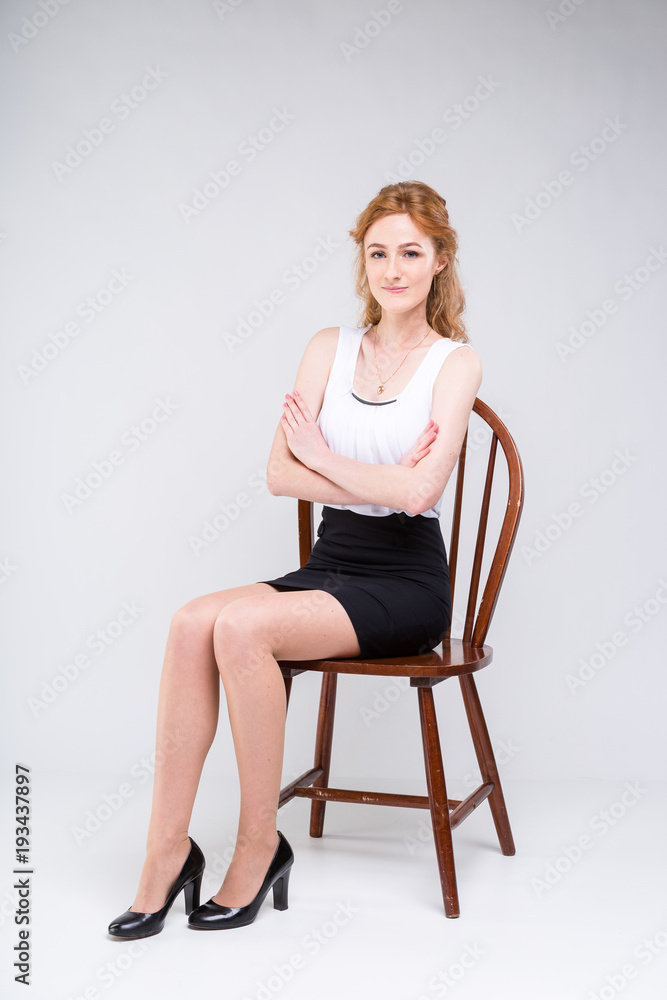 Portrait of beautiful business woman with long red, curly hair sitting on wooden chair on white background in studio. Dressed in a white blouse with a short sleeve and black short skirt and high heels Portrait of beautiful business woman with long red, curly hair sitting on wooden chair on white background in studio. Dressed in a white blouse with a short sleeve and black short skirt and high heels