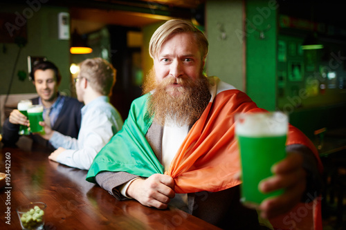 Bearded man with national irish flag and glass of beer looking at camera in pub