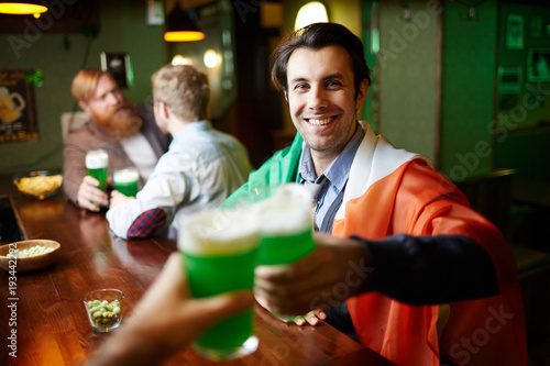 Smiling young man toasting with beer on background of his friends in pub