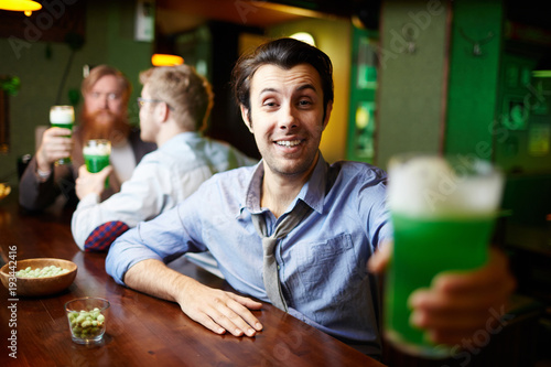 Drunk young man toasting with glass of green beer on background of his friends or companions in pub