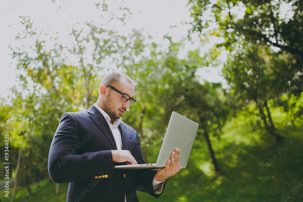 Young successful smart businessman in white shirt, classic suit, glasses. Man standing and working on laptop pc computer in city park outdoors on nature background. Mobile Office, business concept. Young successful smart businessman in white shirt, classic suit, glasses. Man standing and working on laptop pc computer in city park outdoors on nature background. Mobile Office, business concept.