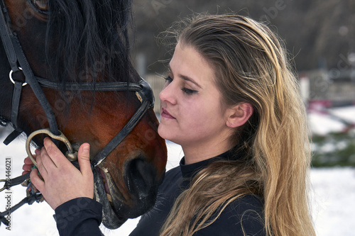 A girl with long blonde hair communicates with her favorite horse. The girl finished riding a horse. A cloudy winter day. Close-up..