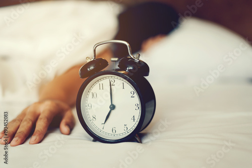 The woman's hand under the blanket reaches out for the alarm clock.