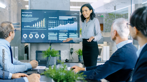 Beautiful Businesswoman Gives Report/ Presentation to Her Business Colleagues in the Conference Room, She Shows Graphics, Pie Charts and Company's Growth on the Wall TV.