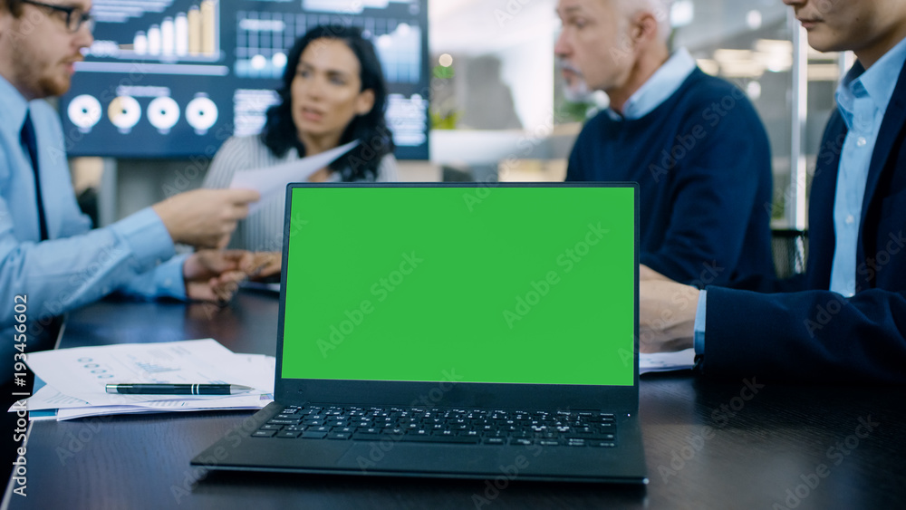 In the Meeting Room Laptop with Green Chroma Key Screen on the Conference Table. In the Background Business People Have Important Discussion. In the Meeting Room Laptop with Green Chroma Key Screen on the Conference Table. In the Background Business People Have Important Discussion.