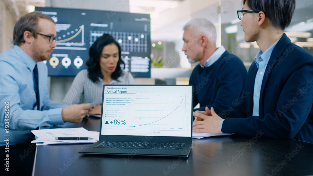 Obraz premium In the Meeting Room Laptop Standing on the Conference Desk Shows Company's Annual Growth Report with Interactive Charts. In the Background Business People Have Important Discussion.