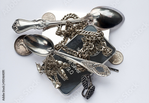 Silver ware are placed on a light gray background. Silver bars, old coins, a teaspoon, a coffee spoon, earrings and chains.