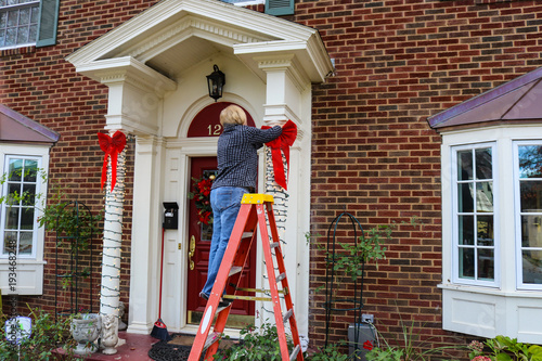 Woman on ladder putting Christmas decorations up on pillars of entrance of two story brick house with bay windows