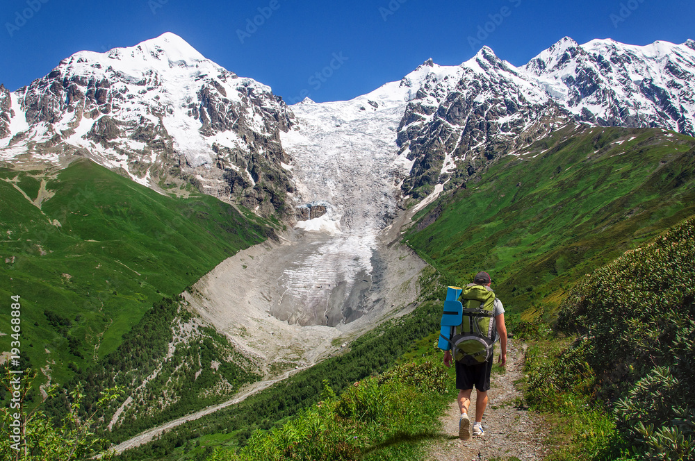 tourist with large backpack rises on mountain trail with view of the Lardaard ice-fall in Georgia Svaneti 