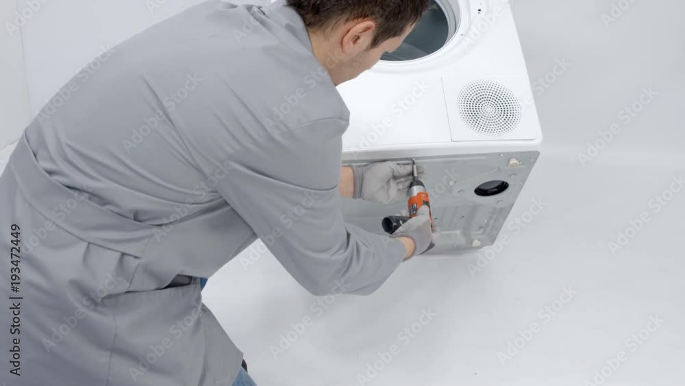 A man is using a drill to take out screws from the back of the washing machine. He needs to fix te device. A man is using a drill to take out screws from the back of the washing machine. He needs to fix te device.