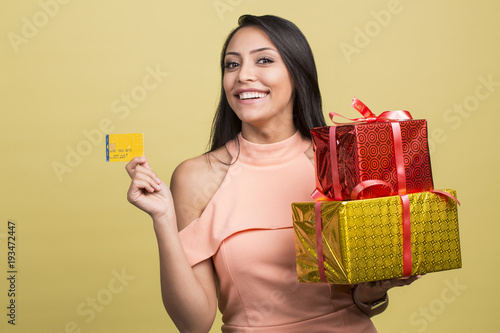 Beautiful young woman holding credit card, present box with gift isolated on yellow background. St. Valentine's Day, Christmas, birthday, shopping concept