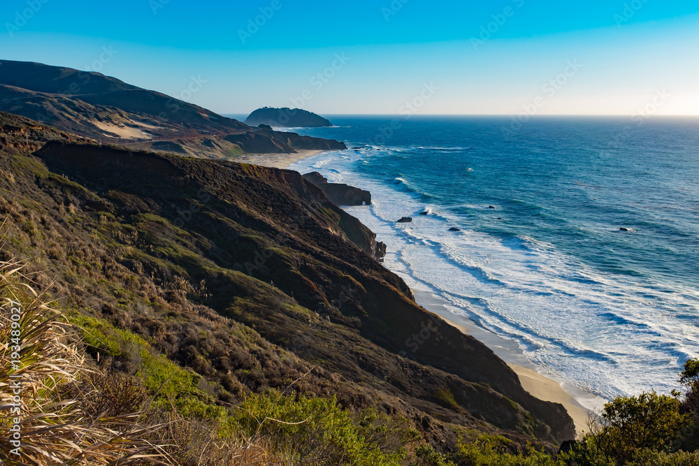 Dramatic landscape view of a Big Sur road trip on a sunny day with waves crushing .