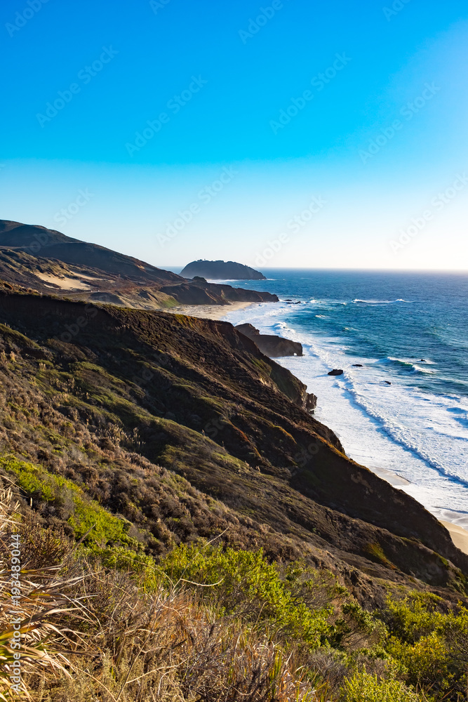 Dramatic landscape view of a Big Sur road trip on a sunny day with waves crushing .