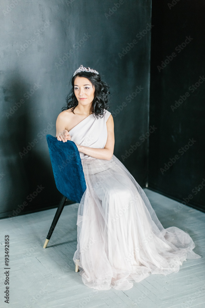 Young beautiful bride with a beautiful diadem in the hair poses sitting on the chair on dark background. Young beautiful bride with a beautiful diadem in the hair poses sitting on the chair on dark background.