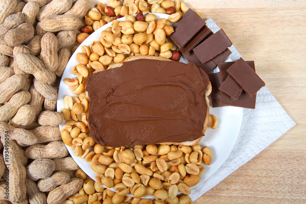 Chocolate peanutbutter spread on bread, open face sandwich on plate with pieces of chocolate, shelled peanuts and whole in shell peanuts on wood table. Tasty combination of chocolate and peanuts Chocolate peanutbutter spread on bread, open face sandwich on plate with pieces of chocolate, shelled peanuts and whole in shell peanuts on wood table. Tasty combination of chocolate and peanuts