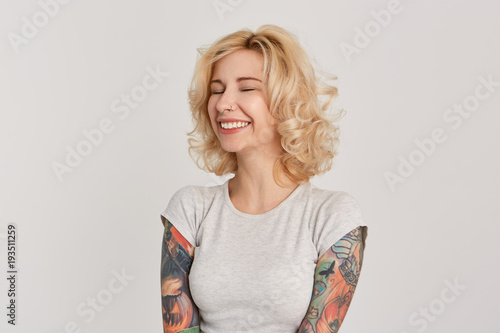 Happiness concept. Portrait of blonde attractive girl, with pierced nose and tattoos over her hands closed her eyes and smiling. Dreaming about birthday gift. Isolated over white wall. photo