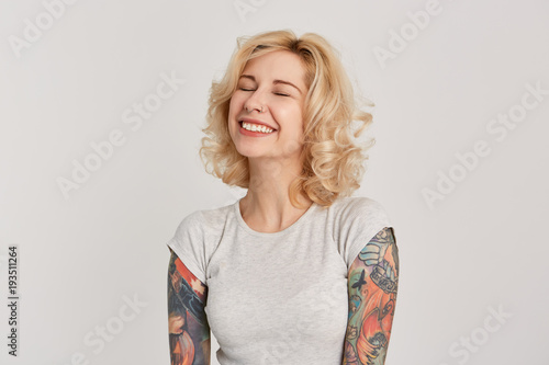 Happiness concept. Portrait of blonde attractive girl, with pierced nose and tattoos over her hands closed her eyes and smiling. Dreaming about birthday gift. Isolated over white wall. photo