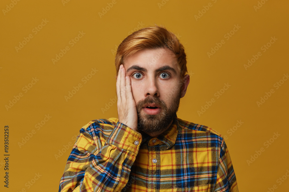 Portrait of stupefied bearded male, with dyed blond gold hair keeps mouth widely opened, wonders about something, isolated over yellow background Portrait of stupefied bearded male, with dyed blond gold hair keeps mouth widely opened, wonders about something, isolated over yellow background