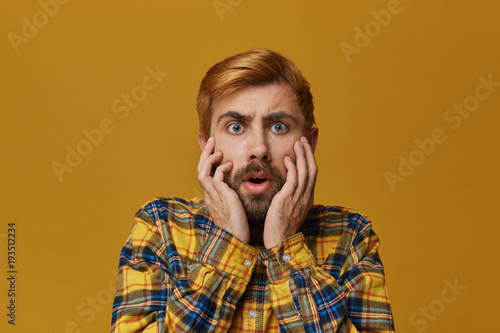 Oh my God! Scared bearded male with dyed hair bieng afraid of something, wathces horror film. Isolated over yellow background