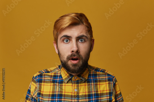 Portrait of stupefied bearded male, with dyed blond gold hair keeps mouth widely opened, wonders about something, isolated over yellow background