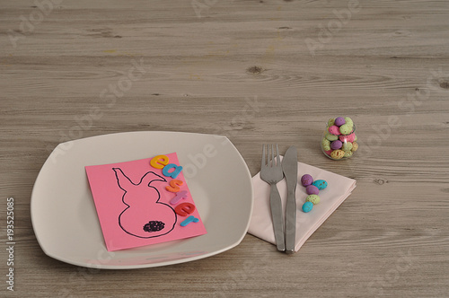 An Easter place setting with a napkin, plate, knife, fork and a bowl of candy displayed on a table with an easter bunny card