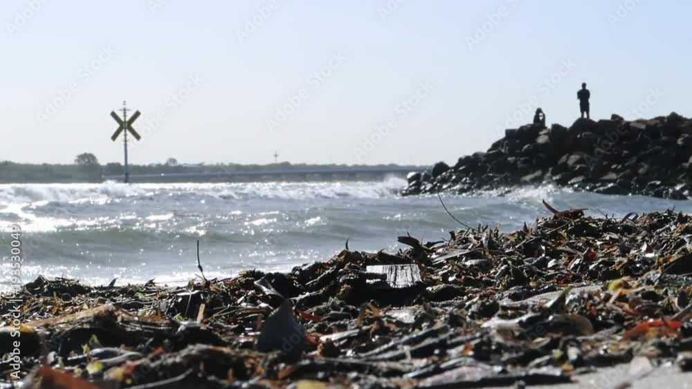 A Ground Level, Long Shot showing the silhouettes of people fishing off a headland. Driftwood in the foreground, bridge in the background and a marine warning sign amongst a rough sea. 