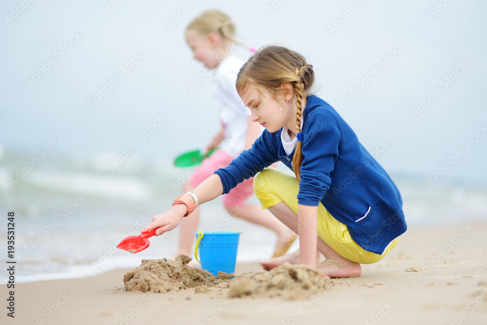 Two little sisters having fun on a sandy beach on warm and sunny summer day. Kids playing by the ocean. Two little sisters having fun on a sandy beach on warm and sunny summer day. Kids playing by the ocean.