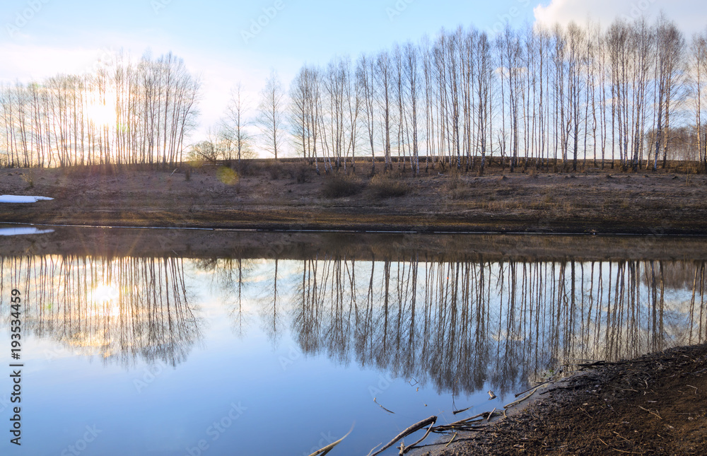 Springtime.Sunny spring colorful landscape with river and bare birches growing on the riverbank.Warm sunlight at sunset.Clouds in blue sky.Beautiful view.Reflection.
