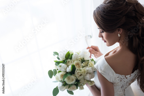 attractive bride in traditional dress with wedding bouquet and glass of champagne standing at window photo