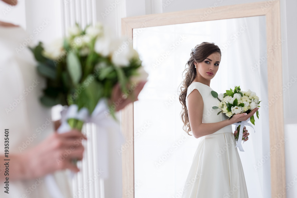 beautiful bride in dress with wedding bouquet looking at her reflection in mirror beautiful bride in dress with wedding bouquet looking at her reflection in mirror