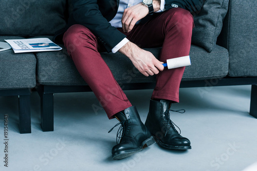 partial view of businessman cleaning pants with sticky brush while sitting on sofa