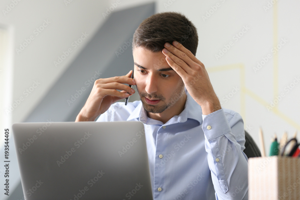 Young man talking on phone while working with laptop in office Young man talking on phone while working with laptop in office
