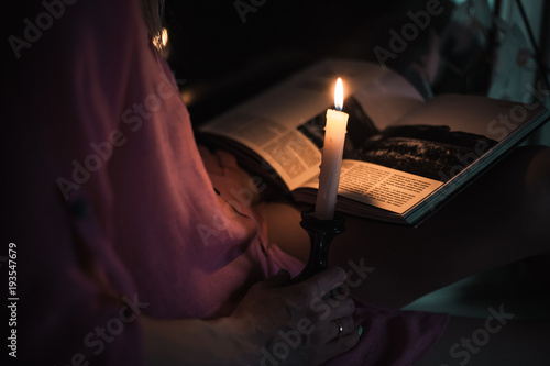 A beautiful young girl in a shirt reading a book at the window-sill by the light of a burning candle at night