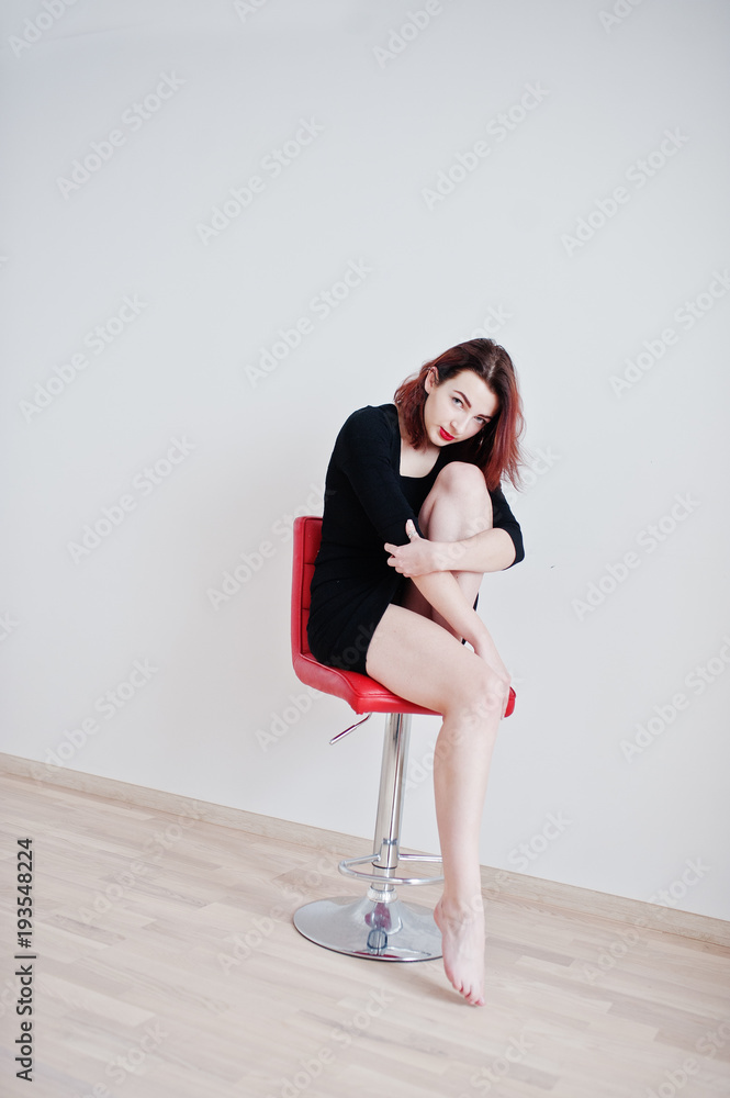 Red haired girl on black dress tunic sitting on red chair against white wall at empty room. Red haired girl on black dress tunic sitting on red chair against white wall at empty room.
