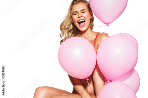 Laughing young woman with pink festive balloons sits on a white background in pink lace underwear screwed up her eyes.