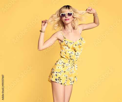 Short-haired girl in Fashionable Sunglasses Dancing. Young Playful female Blond model in Stylish fashion Summer Outfit. Beautiful Happy woman Having Fun dance in Studio on Yellow