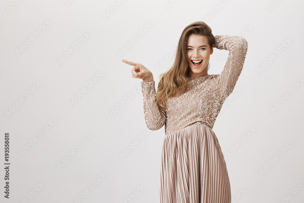 Portrait of playful charming caucasian female in glittering luxurious evening dress holding hand on head and pointing left with index finger, expressing joy and amazement while over gray background. Portrait of playful charming caucasian female in glittering luxurious evening dress holding hand on head and pointing left with index finger, expressing joy and amazement while over gray background.