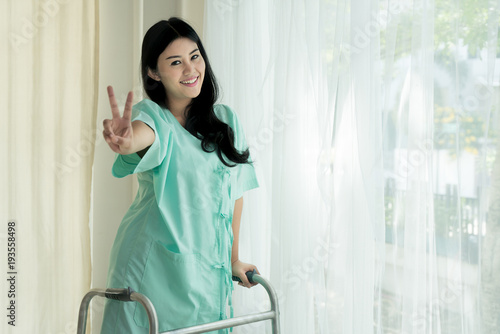Young Asian patient woman standing with Folding walker at hospital room showing victory sign for cheerful.