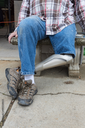 Leg amputee sitting on bench with hands by sides, legs crossed, copy space, vertical aspect © Natalie Schorr