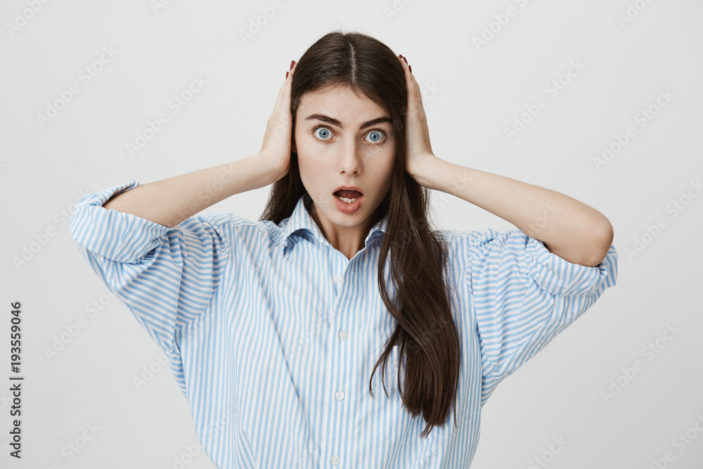 Portrait of attractive woman with stunned and shocked expression, covering her ears with both hands and standing with opened mouth over gray background. Friend shared secret she did not want to know