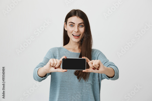 Attractive and smart brunette smiling broadly while making advertisement of new smartphone, holding it with both hands ahead and showing gadget to camera, standing over gray background