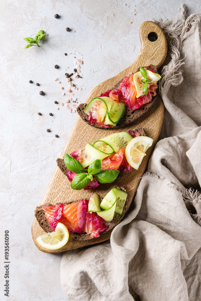 Sliced beetroot marinated salmon sandwiches with rye bread, cucumber, basil and lemon served on wooden cutting board over grey texture background with textile linen. Top view, space