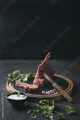 Grilled sliced rack of lamb with yogurt mint sauce served with green salad young beetroot leaves, knife and pink salt on round wooden slate board over dark black table. Copy space. Toned image