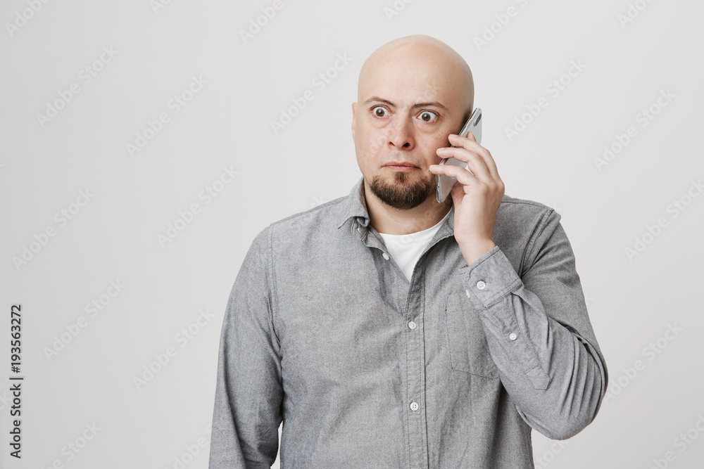 Portrait of amazed worried caucasian bald man talking on smartphone while looking aside with popped eyes and anxious expression, standing over gray background. Someone blackmails him. Portrait of amazed worried caucasian bald man talking on smartphone while looking aside with popped eyes and anxious expression, standing over gray background. Someone blackmails him.